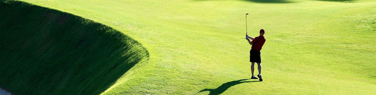 A golfer in a red shirt and black shorts stands on a green golf course, holding a golf club near the hole flag, with bright sunlight casting long shadows across the grass.